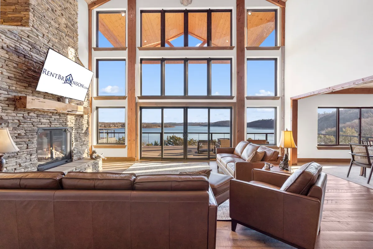 This image showcases the main living area with large windows offering stunning lake views, a striking stone fireplace, comfortable leather sofas, and a TV mounted on the wall. The space is filled with natural light and designed for relaxation and social gatherings.