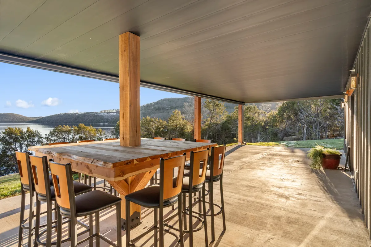 An inviting outdoor wooden table with bar stools overlooking a serene lake, framed by a mountainous backdrop. The scene hints at relaxed social gatherings, morning coffee, or evening drinks with picturesque views.