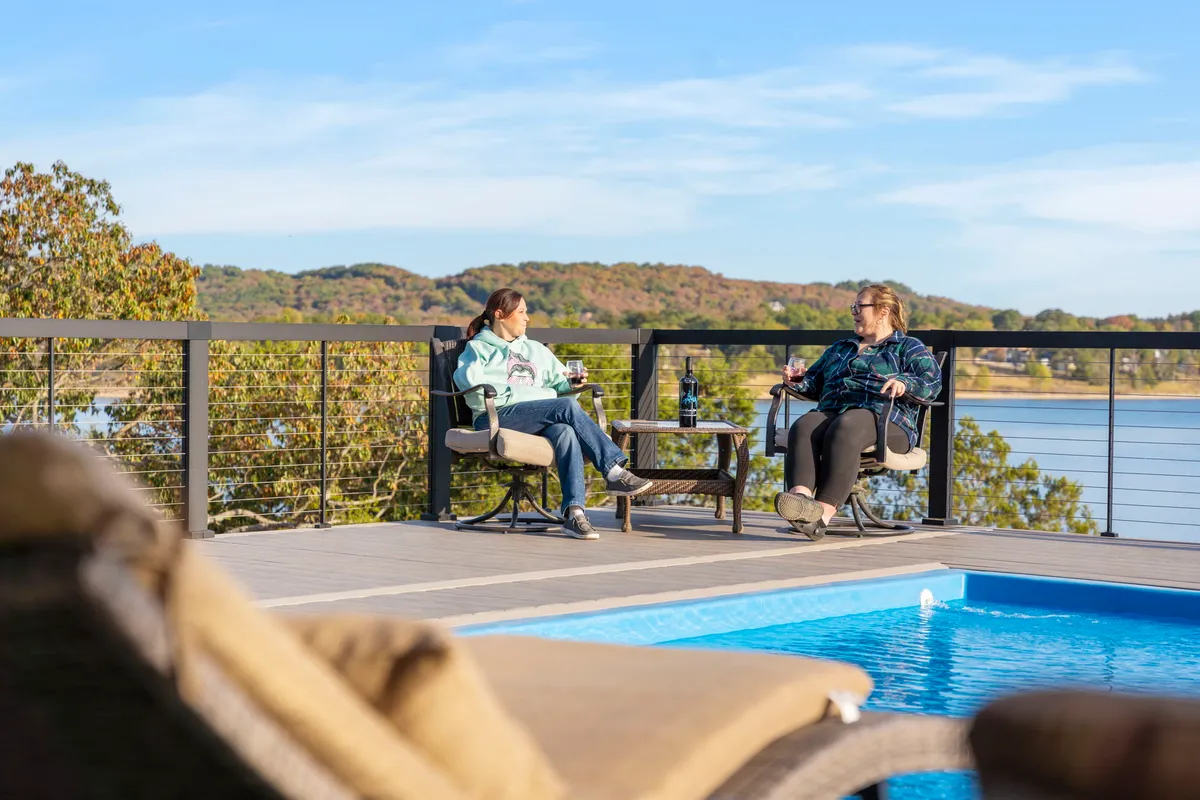 Two women sit comfortably by the pool, drinks in hand, enjoying a peaceful conversation as the lake stretches out in the background. The scene feels casual yet exclusive, emphasizing the poolside social atmosphere and the inviting water.