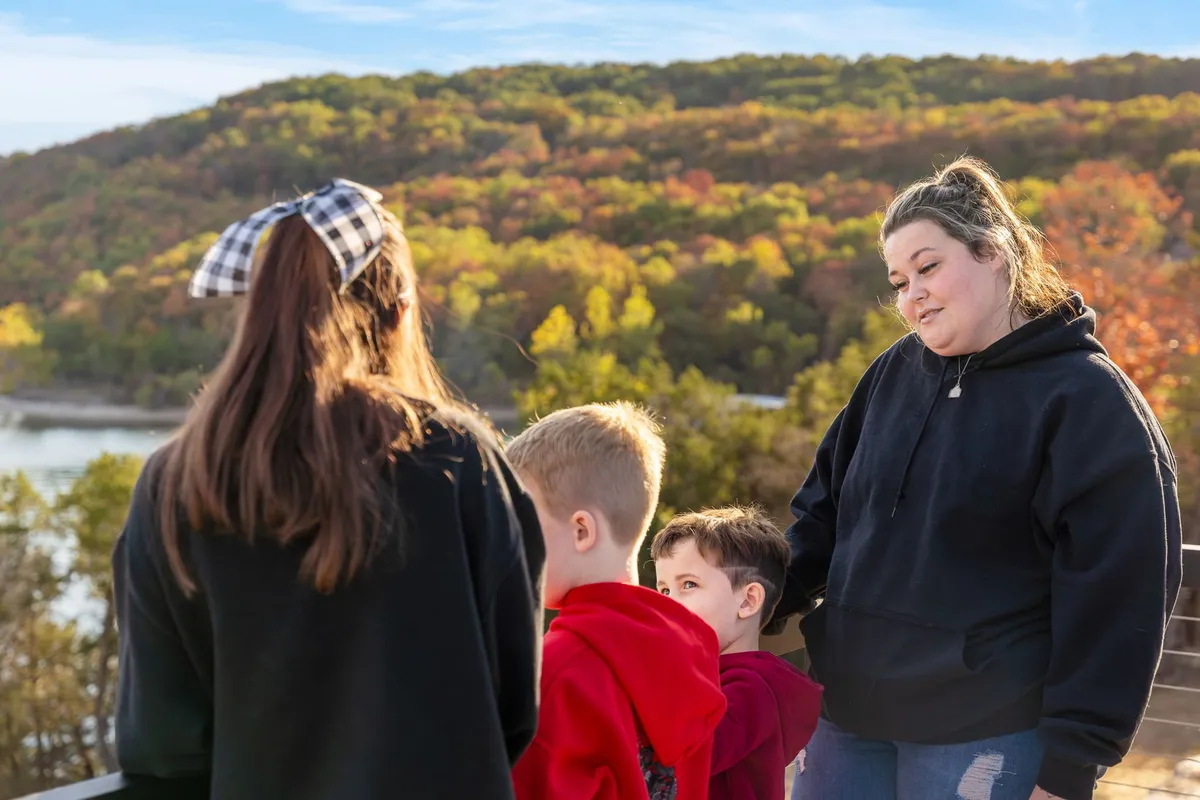 A joyful family scene on a riverside deck, surrounded by vibrant autumn foliage. The family members are engaged in conversation, laughter, and enjoying the natural setting as the sun sets over distant hills.