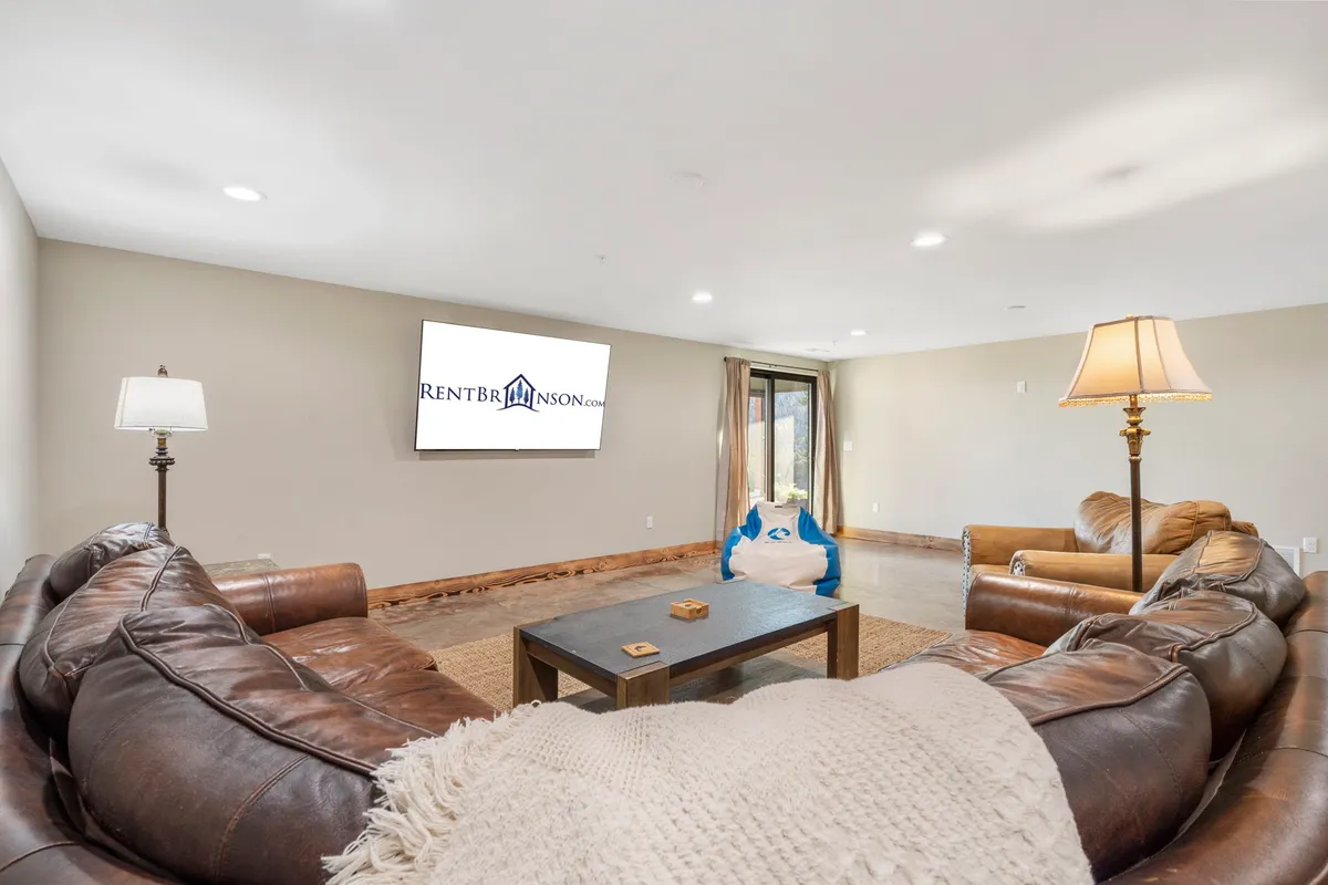 A cozy lounge area featuring brown leather sofas arranged around a large television and coffee table, with soft lighting from a floor lamp. The space is inviting for family gatherings, watching movies, or hosting game nights, echoing the section’s emphasis on relaxation and entertainment on the lower level.