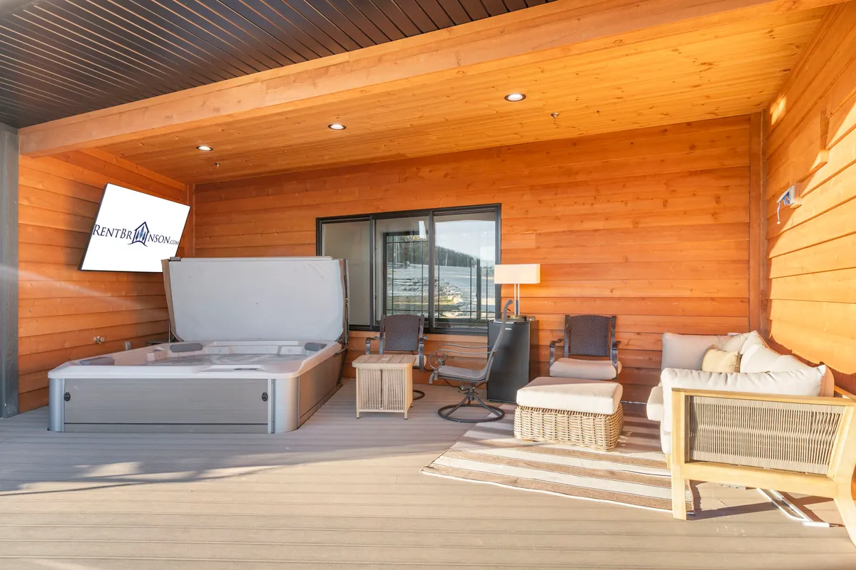 A bubbling hot tub sits on a covered patio, with a cozy seating area facing a 75" outdoor Smart TV. The wooden walls add warmth, and open views to the outdoors suggest lake scenery just beyond.