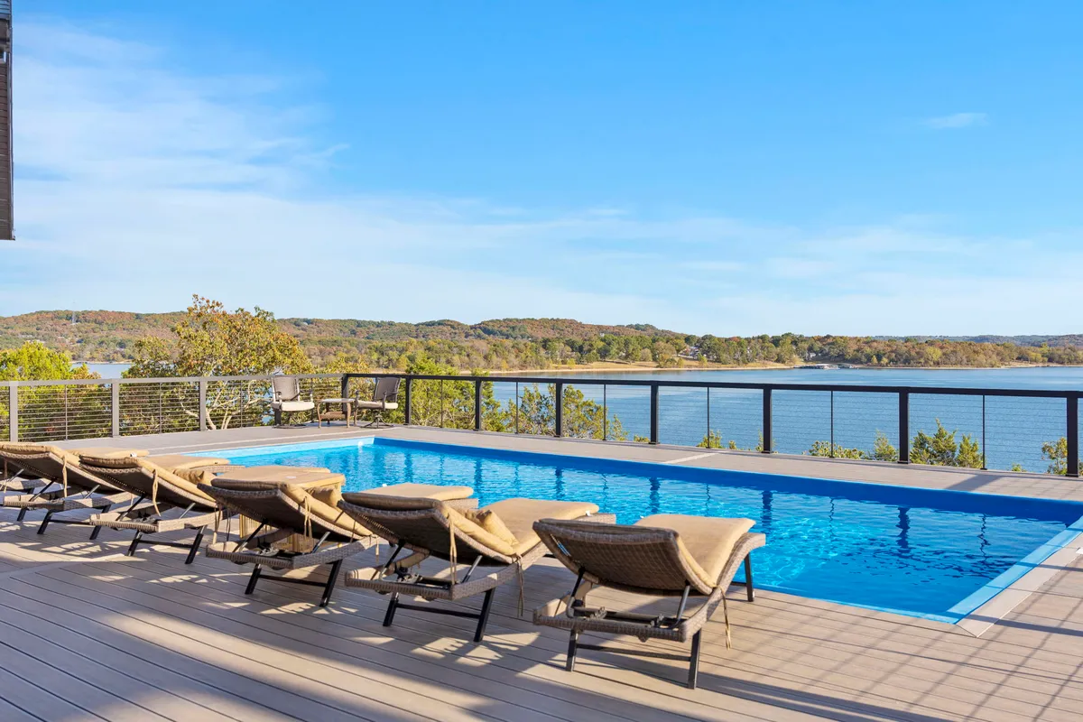This image features a tranquil swimming pool with inviting lounge chairs, all set against a scenic backdrop of a lake and deck railing. The space exudes relaxation, combining the sparkling water, comfortable seating, and a panoramic lake view.