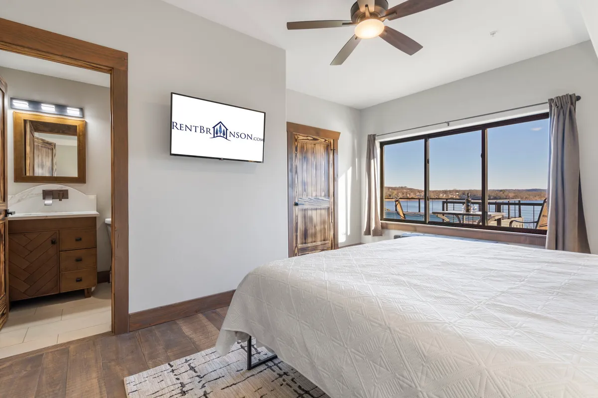 A king bedroom with crisp white bedding, large window offering a serene lake view, a wall-mounted television, and warm wooden furniture. Natural light fills the space, and the décor feels calm and inviting, reflecting a peaceful retreat.
