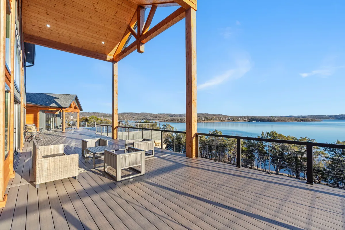 An expansive wooden deck with stylish outdoor furniture overlooks Table Rock Lake, framed by wooden beams. The scene is serene and welcoming, ideal for relaxing, socializing, and enjoying the sunset over the water.