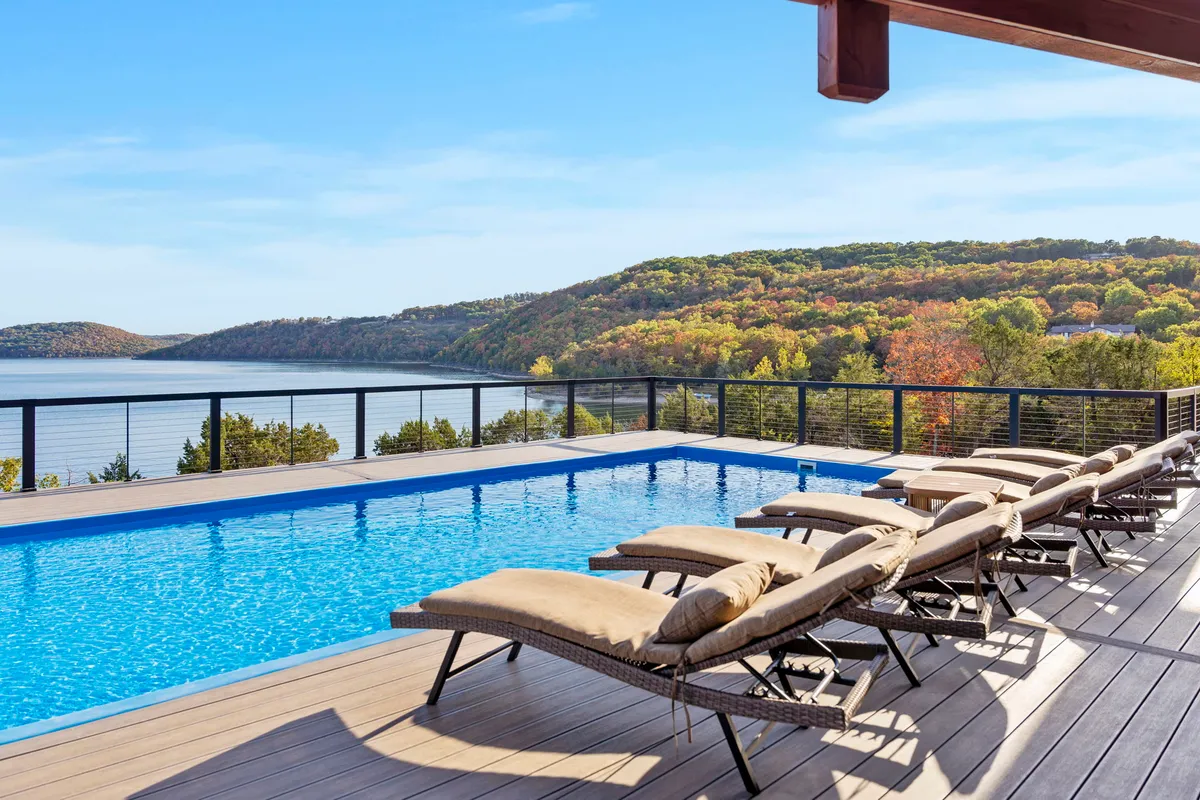 The image showcases the exterior of a newly built lakeside house in Lampe, Missouri, featuring a private swimming pool surrounded by sun loungers. Beyond the pool, there are sweeping, unobstructed views of Table Rock Lake and the rolling hills. The scene captures the golden light of sunset, creating a tranquil, inviting atmosphere perfect for relaxing, socializing, and enjoying the beauty of the outdoors.