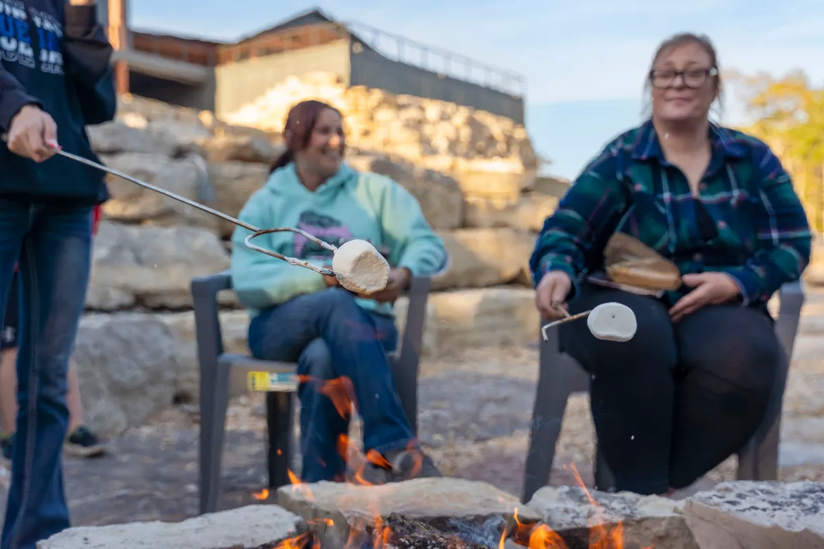 A group gathers around a crackling campfire, roasting marshmallows over glowing embers with rocks forming a rustic fire pit. The scene is joyful and relaxed, with smiles and animated conversation hinting at storytelling and laughter under the stars.