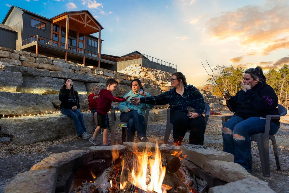 A lively fire pit scene with people interacting, stone seating, and a campfire in the foreground. The house is visible in the background, suggesting proximity to the main outdoor living areas.