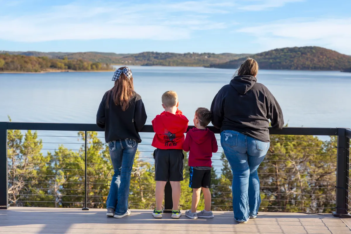 A family stands together overlooking the calm surface of Table Rock Lake, surrounded by distant hills and a lush treeline. The soft evening light suggests the closing of an adventurous day, and the group appears to be enjoying a peaceful, shared moment by the water.