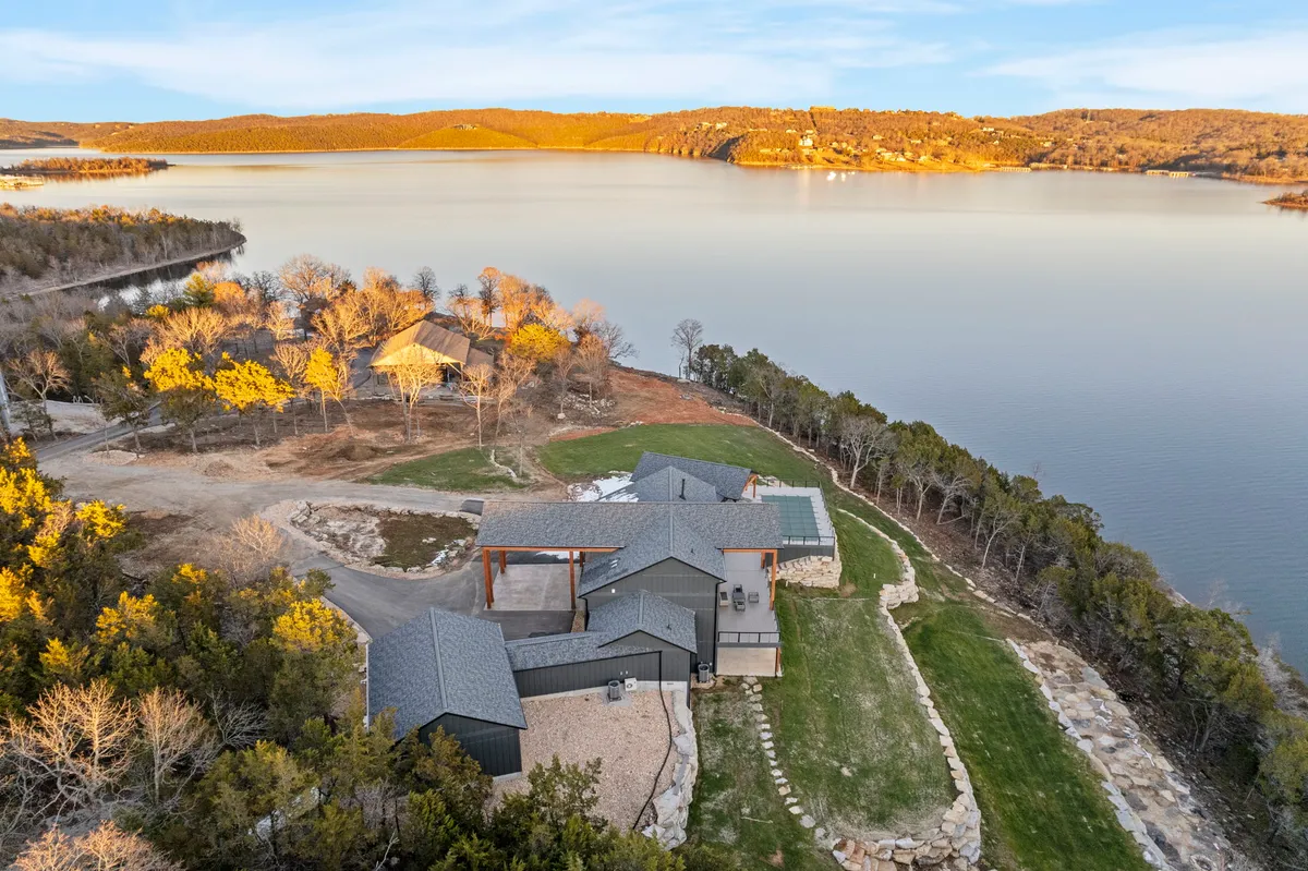 A sweeping view of a tranquil lake bordered by green landscape, with the modern architecture of a lakeside home bathed in a warm sunset glow. The scene hints at opportunities for outdoor activities such as boating, fishing, picnicking, and hiking, with nature trails visible around the property.