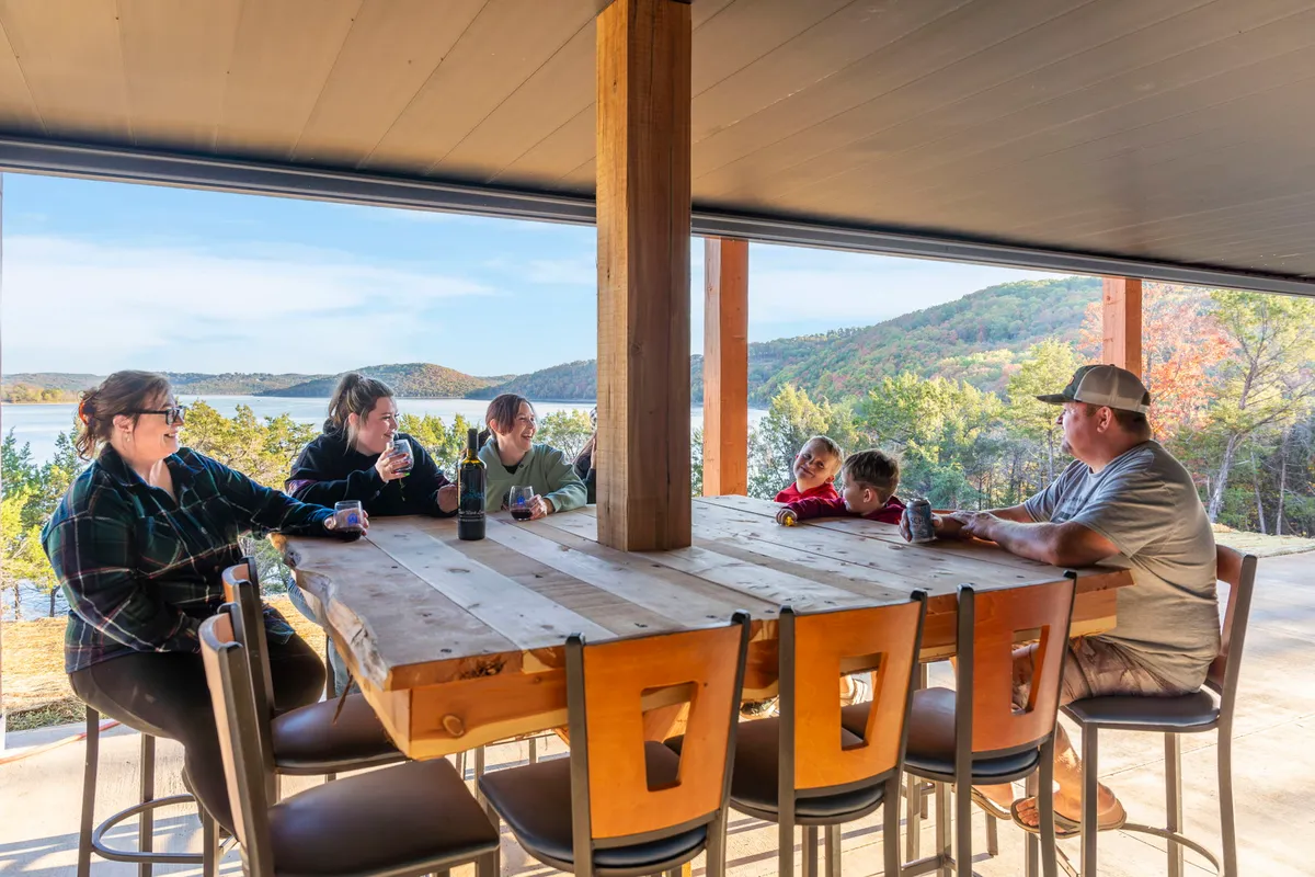 A large wooden outdoor table surrounded by a family enjoying drinks and conversation, set against a backdrop of the lake. This scene captures the covered patio or outdoor dining experience, blending the joy of group gatherings with direct access to the lake and yard.
